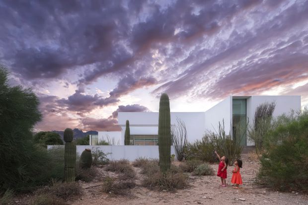 Exterior view of a minimalist white modern residence set within a desert landscape, framed by tall columnar cacti, scrub vegetation, and sandy ground. The building has clean geometric volumes, flat rooflines, large glazed openings, and pale rendered walls. Two people stand in the foreground, while a dramatic purple and pink sunset sky forms the backdrop.