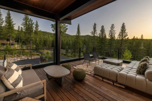 A spacious covered outdoor terrace overlooking a forested landscape. The terrace features warm timber decking and ceiling lining, dark structural framing, clear glass balustrades, a large cream modular sectional sofa, lounge chairs, a low coffee table, and decorative outdoor seating. The setting feels refined, warm, and connected to nature.