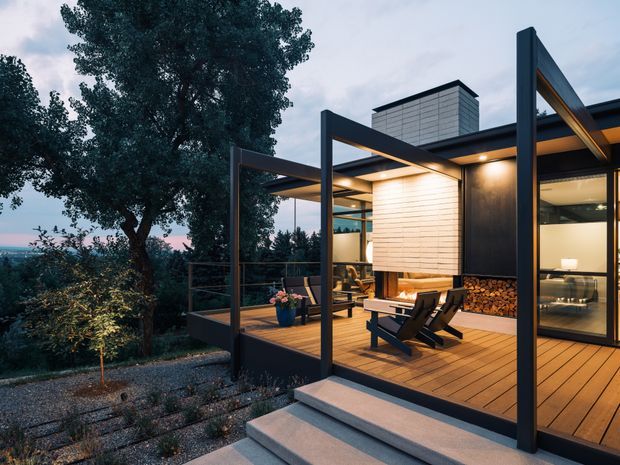 Twilight exterior view of a contemporary elevated pavilion-style home with extensive glazing, dark structural steel framing, warm timber decking, and a covered outdoor terrace. The terrace includes lounge chairs, a built-in firewood storage feature, and warm integrated lighting. Steps connect the deck to a landscaped garden, with mature trees and a distant water or coastal outlook visible beyond.