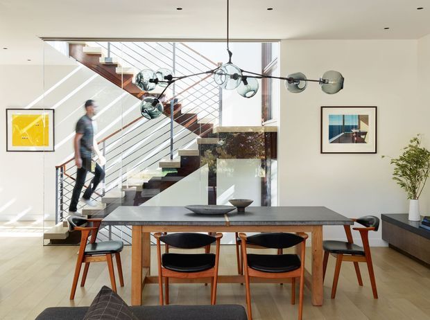 Bright, open-plan dining area with a mid-century-inspired timber dining table and sculptural chairs, positioned in front of a dramatic open staircase. The room features pale timber flooring, white walls and ceiling, a large glass stair balustrade, black metal stair detailing, framed artwork, a low dark sideboard, and a distinctive multi-globe pendant light. A living area is partially visible in the foreground.