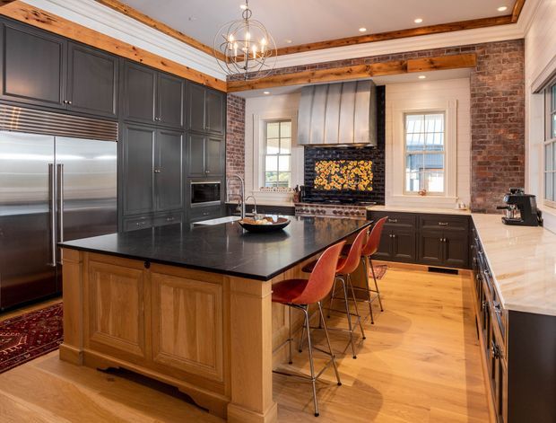 A spacious, richly detailed kitchen with a large central timber island topped in black stone and surrounded by red bar stools. The room features dark charcoal cabinetry, stainless-steel appliances, a pale stone perimeter benchtop, exposed timber ceiling beams, painted ceiling mouldings, exposed red brick walls, timber flooring, and a decorative metal rangehood above a patterned tiled or mosaic cooking backsplash. Multiple windows provide natural light, complemented by recessed ceiling lights and a sculptural pendant.
