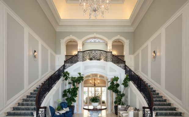 Grand formal residential entry hall with a symmetrical split staircase, ornate dark metal balustrades, white architectural mouldings, arched openings, paneled pale grey walls, polished light flooring, and a large decorative chandelier. The space has a double-height central void and an elegant traditional character.