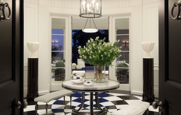 A refined formal entry foyer viewed through open black double doors. The bright, curved bay window is framed by white fluted wall panelling and French doors, while a black-and-white checkerboard floor anchors a round metal console table with a large floral arrangement. Sculptural black pedestal stands, white decorative vessels, upholstered benches, and a contemporary chandelier create a polished, high-contrast interior.