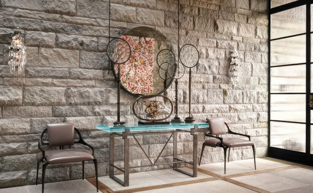 A refined entryway or reception area featuring a dramatic exposed stone block wall, a glass console table with metal supports, sculptural circular metal artwork, contemporary upholstered chairs, crystal wall sconces, and a large black-framed glazed door or screen. The composition has a luxurious, gallery-like feel with strong textural contrast between the masonry, glass, and metal.