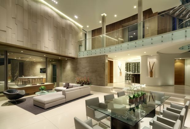 A spacious, sophisticated open-plan interior with a double-height living and dining area. The room features a large pale sectional sofa, glass dining table with upholstered chairs, sculptural lounge seating, polished light flooring, a textured stone or concrete feature wall, extensive timber wall panelling, recessed lighting, and a glazed mezzanine-level balustrade. A kitchen or bar area is visible beyond the living space.