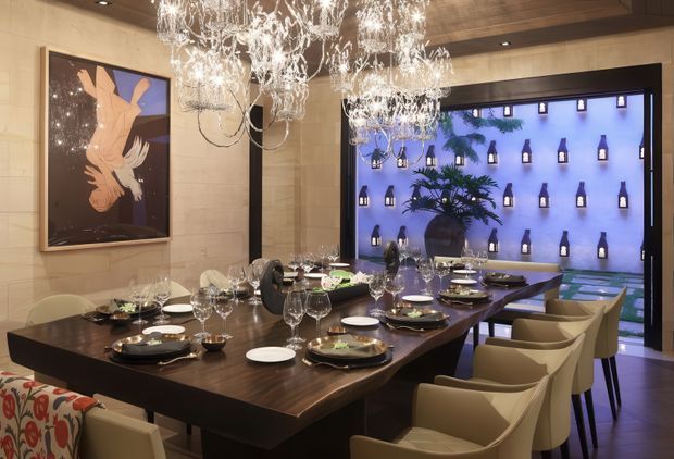 Luxurious contemporary dining room with a long dark wood dining table set for a large gathering, surrounded by upholstered chairs. Multiple ornate crystal chandeliers hang beneath a dark timber ceiling, while a stone-clad wall, framed artwork, and a dramatic blue-lit bottle display visible through glazing create a sophisticated restaurant-like atmosphere.