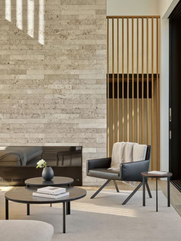 Contemporary living room with a tall textured stone-clad feature wall, black built-in media joinery, and a timber vertical-slat screen creating a partial double-height feel. The room is furnished with a black leather lounge chair, round nesting coffee tables, neutral textiles, and subtle decor. Warm sunlight and neutral materials create a refined, calm atmosphere.