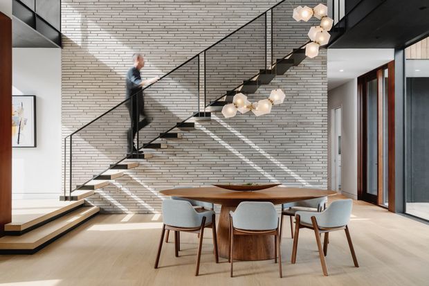 A bright contemporary open-plan dining area beneath a dramatic zigzag staircase. The space features a pale timber floor, a large sculptural timber dining table with upholstered chairs, a full-height light brick feature wall, slender black metal stair rails, and clustered pendant lighting. Dark built-in cabinetry and tall glazed doors frame the edges of the room.