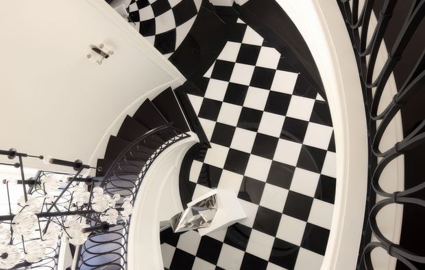 A dramatic overhead view of a sweeping curved stairway and landing with ornate black metal balustrades, white walls, and a striking black-and-white checkerboard floor. A sculptural chandelier hangs below, reinforcing the grand, theatrical character of the interior.