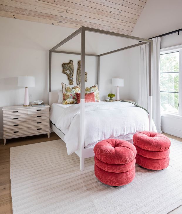 Bright bedroom with a white four-poster canopy bed, crisp white bedding, pale timber vaulted ceiling, neutral walls, and a light striped rug. A timber dresser and matching bedside lamps sit to the left, while two vivid coral-red tufted ottomans provide a bold accent at the foot of the bed. Tall windows with white curtains bring in abundant natural light.