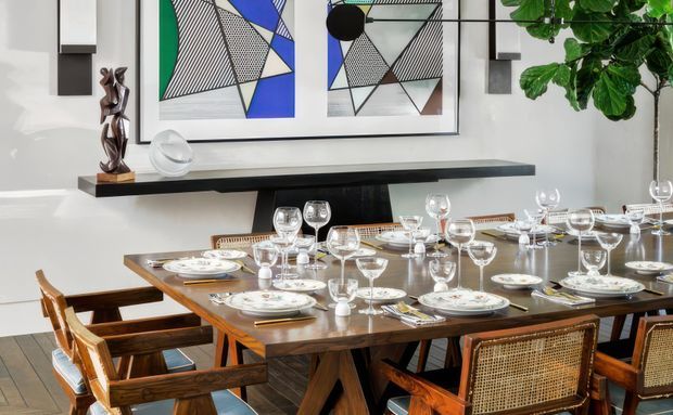 A refined dining room with a large timber dining table formally set with white crockery and numerous wine and cocktail glasses. Wooden dining chairs include woven cane backs, while a dark floating console shelf, sculptural decor, geometric abstract artwork, wall sconces, and a large leafy indoor plant complete the space.