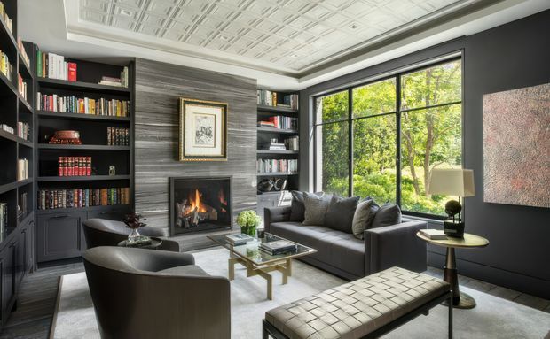 Sophisticated dark-toned living room library with floor-to-ceiling built-in bookshelves, a stone-clad fireplace, large black-framed windows overlooking greenery, a grey sofa and lounge chairs, a glass coffee table, and a tufted leather bench. The room has a decorative pressed-tin ceiling and a refined, luxurious atmosphere.