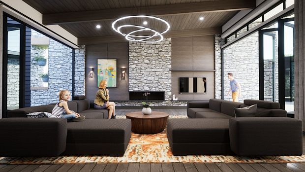 A spacious contemporary living room with multiple dark modular sofas arranged around a round wooden coffee table. The room features floor-to-ceiling black-framed glazing, a dramatic rough stacked-stone fireplace wall, warm wood ceiling and flooring, integrated timber-toned cabinetry, and sculptural ring pendant lighting. A patterned rug adds colour beneath the seating area.
