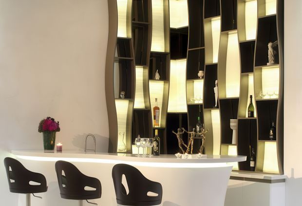 A sophisticated contemporary home bar or entertaining counter with a sculptural curved white island and three dark bar stools. Behind it is a full-height black built-in shelving wall with irregular geometric compartments, illuminated translucent panels, glassware, bottles, and decorative objects. The space has warm integrated lighting and a refined, dramatic atmosphere.