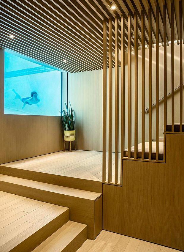 A warm, contemporary interior stairway with light timber flooring, stepped platforms, and a sculptural vertical timber-slat balustrade extending into a slatted ceiling. A large illuminated underwater pool image or window forms a vivid blue feature on the wall, complemented by a potted plant and integrated recessed lighting.