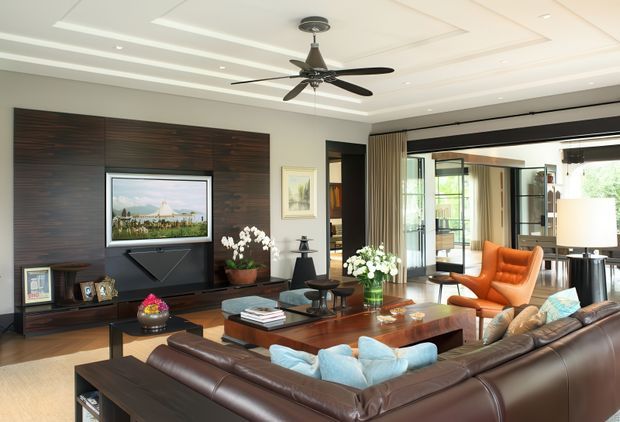 A spacious contemporary living room with a dark timber feature media wall, wall-mounted television, layered recessed ceiling detailing, ceiling fan, large brown leather sectional sofa, timber coffee tables, and a bright orange lounge chair. Full-height glazing and sliding doors open the room toward an outdoor area, while decorative plants and artwork add softness.