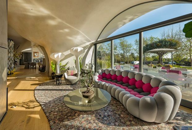 A sculptural, light-filled living room with a curved cream ceiling and expansive floor-to-ceiling glazing overlooking a landscaped outdoor area. The room features a distinctive rounded modular sofa in grey with vivid pink cushions, rounded sculptural lounge chairs, a circular glass coffee table, patterned area rugs, timber flooring, indoor plants, and an open connection to a dining or outdoor entertaining area.