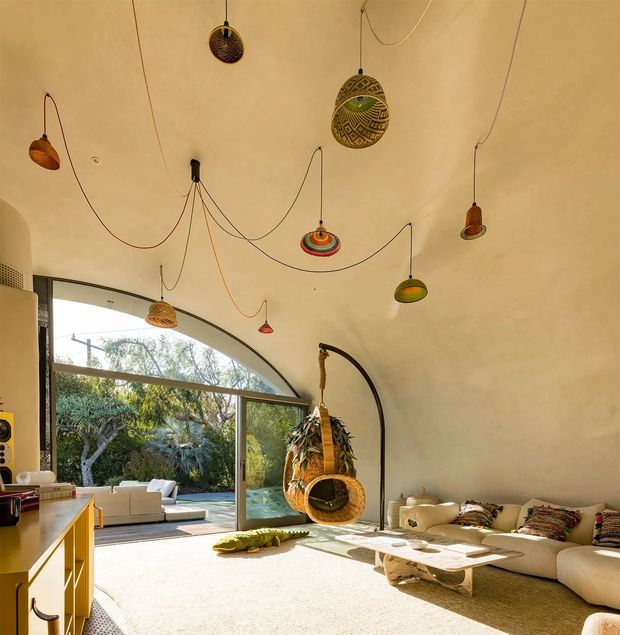Sunlit eclectic living room with a dramatic curved glazed wall opening onto a landscaped outdoor area. The room features a cream curved sectional sofa, low organic coffee tables, a hanging woven egg chair, colourful handmade pendant lights suspended from exposed cords, and warm neutral plaster surfaces. A compact yellow built-in cabinet and partial kitchen joinery are visible at left. The space has a playful, bohemian character with strong indoor-outdoor connection.