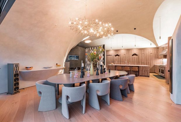 A sophisticated open-plan dining and kitchen interior with a large oval dining table surrounded by upholstered sculptural chairs. The room features warm timber flooring, extensive vertical timber wall panelling and cabinetry, a curved sculptural ceiling, a dramatic branching chandelier, and a dark stone or tiled feature wall behind the dining area. The kitchen occupies the rear of the space with a long island, pendant lights, integrated cabinetry and appliances. The overall composition is warm, luxurious and highly designed.