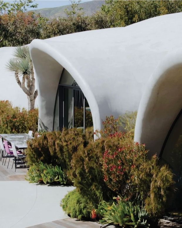 Exterior view of a sculptural, single-storey building with smooth white curved walls and a flowing flat/organic roof form. Dark-framed floor-to-ceiling glazing is set into the arched openings. The building is surrounded by drought-tolerant landscaping, succulents and desert-style planting, with an outdoor dining area visible to the left and mountainous terrain in the background.