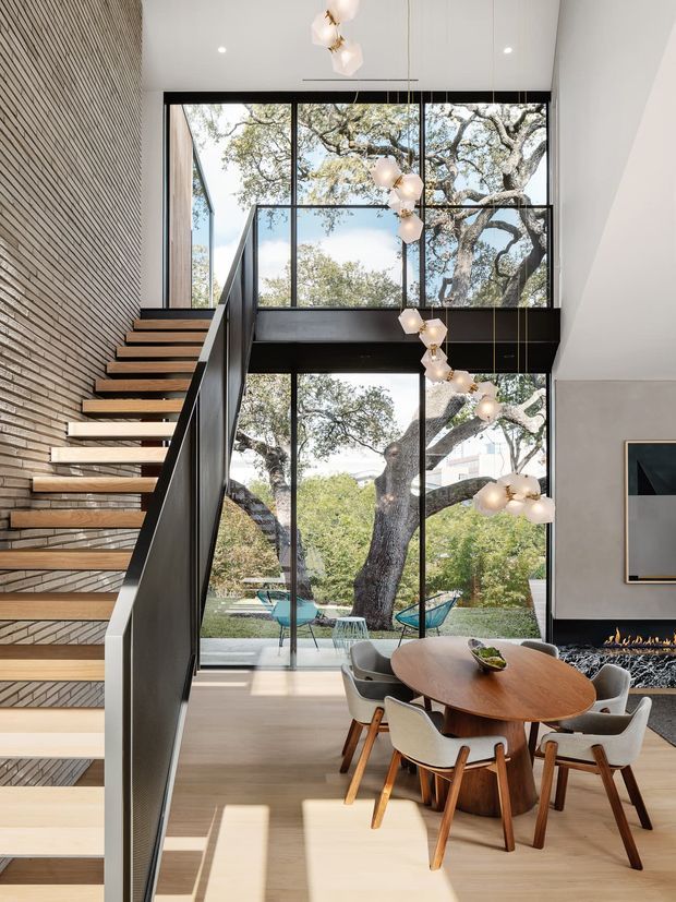 Bright contemporary dining area beneath a dramatic double-height glazed void, with a floating timber staircase and black metal balustrade beside a textured brick wall. Full-height black-framed glazing overlooks mature trees and an outdoor seating area. A sculptural pendant installation hangs above a rounded timber dining table with upholstered timber chairs, while a linear fireplace and artwork are visible to the right.