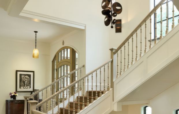 A bright, refined residential stair hall with a sweeping timber staircase, painted balustrade and turned spindles, arched multi-pane windows, decorative wall mouldings, and a pendant light. A console table and framed artwork sit beneath the staircase, while sculptural wall art adds a contemporary accent.