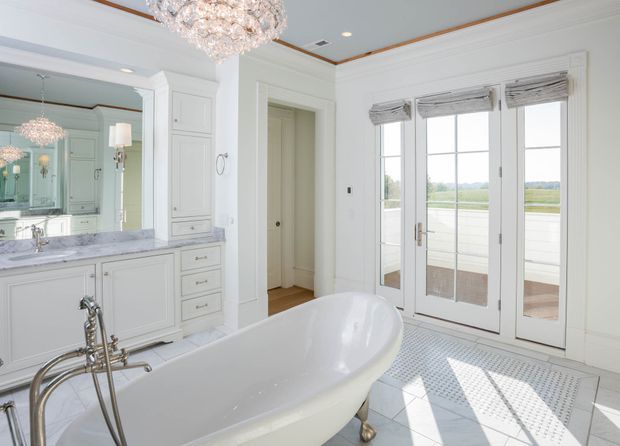 Bright, elegant bathroom featuring a freestanding white bathtub with polished metal feet and traditional tapware, a double vanity with pale cabinetry and a marble-look benchtop, a large wall mirror, ornate crystal chandelier, white decorative wall mouldings, and full-height French doors opening to a balcony or terrace. The room has abundant natural light and a refined classic aesthetic.