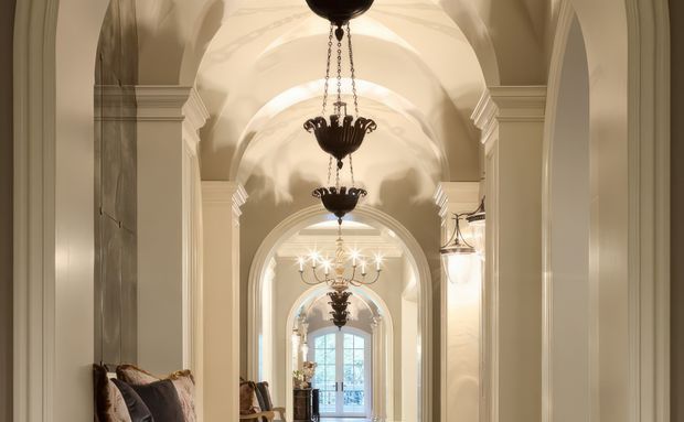 A long, elegant interior corridor framed by repeated rounded arches and pilasters, with a sequence of ornate dark chandeliers suspended from a richly moulded barrel-vaulted ceiling. Warm cream plaster, classical detailing, wall sconces, and glimpses of seating create a refined, grand atmosphere, with daylight visible through glazed doors at the far end.