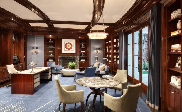 A richly detailed traditional study-library and sitting room with dark timber wall panelling, extensive built-in bookshelves, a coffered and panelled ceiling, blue carpet, a substantial desk, lounge seating, and a round marble-topped table. Tall glazed doors open to an exterior terrace or garden, while warm integrated lighting and classic furnishings create a refined, luxurious atmosphere.