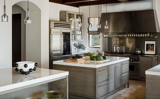 A refined contemporary kitchen with a large central island, pale stone countertops, grey wood-look cabinetry, stainless-steel appliances, dark bronze or black splashback and rangehood, exposed dark ceiling beams, and multiple glass pendant lights. An arched opening connects the kitchen to an adjacent space, while warm tiled flooring and layered lighting create a polished, inviting atmosphere.