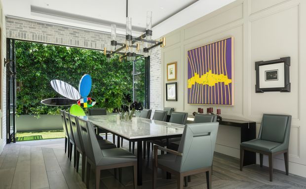 A bright, refined dining room with a long rectangular dining table surrounded by upholstered chairs. The room features extensive white painted wall panelling, pale timber flooring, a large black-framed opening to a leafy garden, a sculptural contemporary chandelier, and colourful abstract artwork. A decorative sculptural object is positioned near the garden opening.