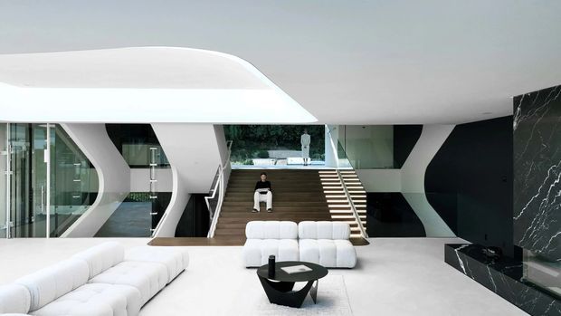 A dramatic, expansive contemporary interior with a sculptural white architectural shell, open-plan lounge, and broad central staircase leading to an upper-level gallery. White modular sofas surround a black geometric coffee table, while dark marble-clad fireplace or feature joinery anchors the right side. Floor-to-ceiling glazing, dark timber stair treads, and strong black-and-white contrast create a luxurious futuristic atmosphere.