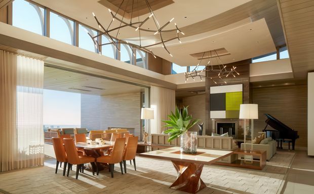A spacious, refined open-plan interior combining a dining area and living room beneath a high sculptural ceiling. Floor-to-ceiling glazing and clerestory windows bring in abundant daylight, while warm timber wall and ceiling finishes, stone or tile flooring, contemporary linear chandeliers, a grand piano, and a geometric feature artwork create a luxurious modern setting. The dining table sits beside the glazed opening, with a lounge area and fireplace or media wall beyond.