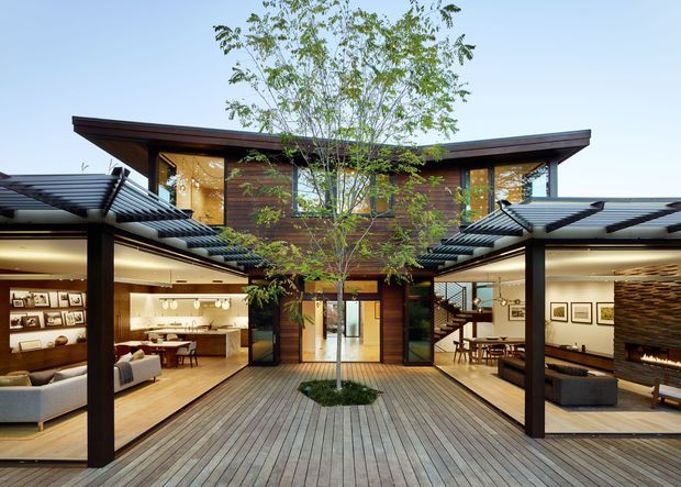 A contemporary multi-wing residence arranged around a central timber deck courtyard with a young tree. The open-sided ground floor reveals warm timber interiors, living and dining areas, framed artwork, and a prominent stone fireplace, while the upper level features dark timber cladding, extensive glazing, and broad projecting roof forms. Black metal pergola structures and exposed structural posts define the courtyard-facing facade.