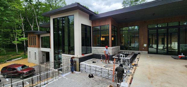 Exterior view of a large contemporary multi-level house under construction in a wooded setting. The building has flat and low-pitched roof planes, extensive black-framed glazing, pale masonry walls, warm timber-clad sections, and a broad covered outdoor terrace. Construction workers and an unfinished pool or retaining structure are visible in the foreground.