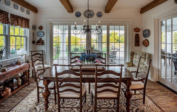 A traditional dining room centred on a large dark timber dining table with ladder-back chairs, a patterned area rug, and a classic chandelier. White painted walls and ceiling are complemented by exposed dark timber ceiling beams, decorative blue-and-white wall plates, and extensive French doors and windows opening toward an outdoor terrace and garden. The room has a warm, collected country-house character with abundant natural light.