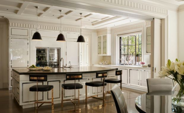 Elegant, spacious traditional kitchen with a large central island and seating for four. The room features detailed white panelled cabinetry, extensive decorative wall and ceiling moulding, dark stone island and benchtops, integrated appliances, pendant lighting, a large divided-pane window, and polished dark timber flooring. A dining area is partially visible in the foreground.