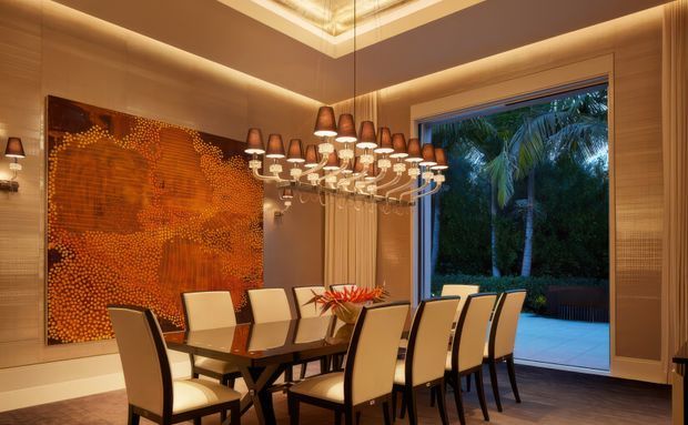 A formal, warmly lit dining room with a long dark dining table and upholstered high-back chairs. A large sculptural chandelier with numerous pendant shades hangs over the table, while a vivid orange abstract artwork forms a feature wall. Full-height glazed doors open to a lush tropical garden and paved outdoor area.