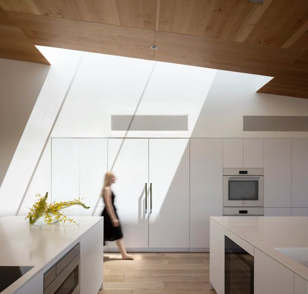 Bright minimalist kitchen with full-height white cabinetry, integrated stainless-steel oven and microwave, and large white island benches. Warm timber ceiling panels and pale timber flooring contrast with the white joinery. Strong sunlight enters through a large overhead skylight or clerestory opening, creating dramatic diagonal shadows across the cabinetry and floor.