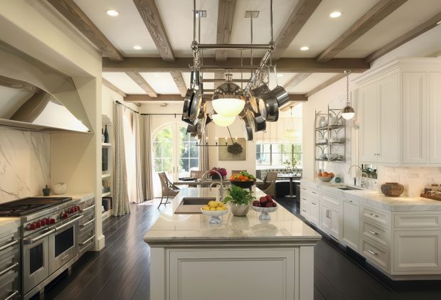 A spacious, traditionally styled kitchen centered on a large marble-topped island. The room features white raised-panel cabinetry, dark timber flooring, exposed rustic wood ceiling beams, a large professional range with hood, and a prominent hanging pot rack over the island. Open shelving, pendant lighting, and tall glazed doors add warmth and brightness.