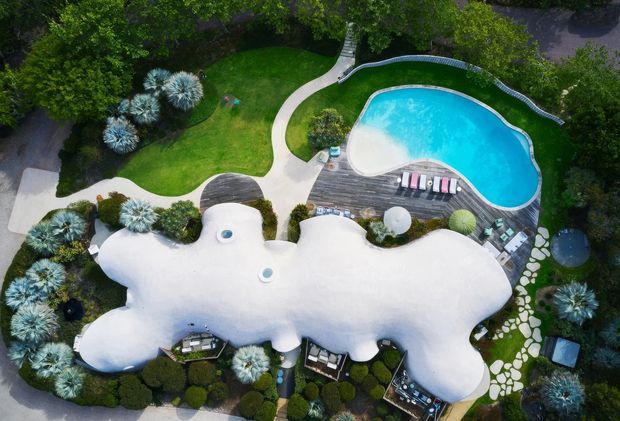 Aerial view of an organically shaped white architectural structure surrounded by landscaped gardens, curved concrete pathways, sculptural plantings, and a bright swimming pool with a timber deck and loungers.