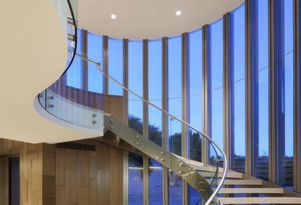 A dramatic contemporary interior featuring a sweeping curved staircase with a sculptural metal stringer, glass balustrade, and timber treads. The staircase rises through a tall, curved glazed facade with slender dark mullions, while warm timber wall panelling and recessed ceiling lights create contrast with the cool blue exterior light. The image emphasises the stairway and the double-height glazed volume.