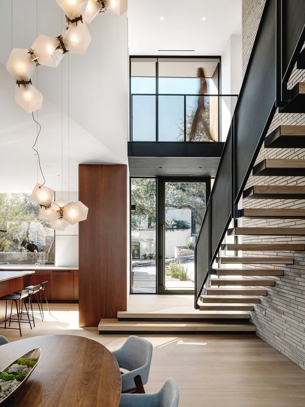 Bright, double-height residential interior combining an open kitchen and dining area with a sculptural floating staircase. The staircase has dark metal framing, open timber treads, and a white brick feature wall. Floor-to-ceiling glazing opens toward a landscaped outdoor area, while a mezzanine-level glazed balustrade overlooks the space. Warm timber cabinetry and a large rounded dining table contrast with the white walls and pale flooring; distinctive faceted pendant lights hang above the kitchen and dining zones.
