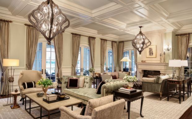 A refined, spacious traditional living room with ornate coffered ceiling, tall draped windows, decorative chandeliers, a carved fireplace mantel, upholstered sofas and armchairs, and layered neutral furnishings arranged around a central coffee table. The room has warm cream, taupe, beige, and light wood tones with patterned textiles and elegant classic detailing.