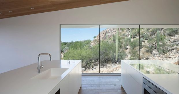Minimal, contemporary kitchen with white island cabinetry and integrated sink, a timber-lined ceiling, pale timber flooring, and expansive floor-to-ceiling glazing overlooking a rugged desert landscape with cacti and rocky terrain. The composition emphasizes clean lines, natural light, and a strong indoor-outdoor connection.