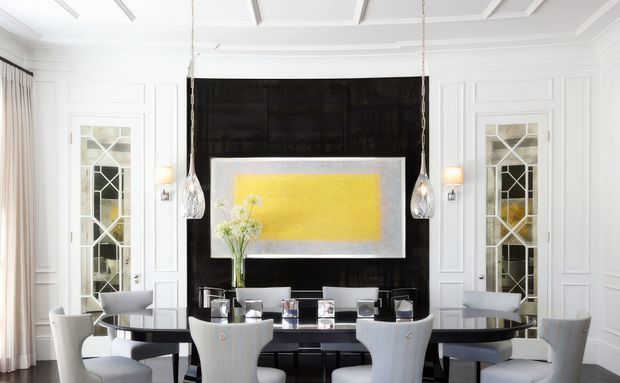Bright formal dining room with a long dark dining table surrounded by upholstered light-grey chairs. White paneled walls, decorative leaded-glass doors, coffered ceiling detailing, pendant lights, and a dramatic black feature wall with a large yellow artwork create a refined, high-contrast setting.