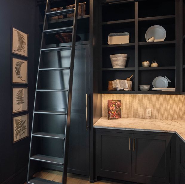A moody, dark-toned built-in kitchen or pantry wall with floor-to-ceiling black cabinetry and open display shelving. A rolling library ladder provides access to upper storage, while framed botanical prints decorate the adjacent wall. The lower cabinets support a light stone countertop and a warm herringbone-patterned backsplash, with dishes and decorative objects arranged on the shelves.