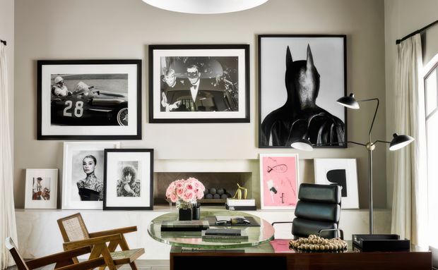 A refined home office or study with a large glass-topped desk, black leather office chair, and sculptural task lighting. The warm neutral room features a marble-look fireplace surround with a recessed dark hearth, tall curtained windows, and an eclectic gallery wall of black-and-white photography, portrait artwork, and graphic prints. The space has a curated, sophisticated feel with layered framed art and dark wood furniture.
