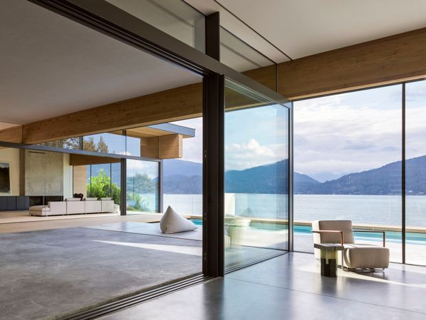 A contemporary open-plan living space with expansive floor-to-ceiling glazing and large sliding glass doors opening onto a waterfront terrace. The room features polished concrete flooring, warm timber beams and wall cladding, dark metal door frames, and restrained modern furnishings. Mountains and water are visible beyond the glazing, creating a bright, airy connection to the landscape.