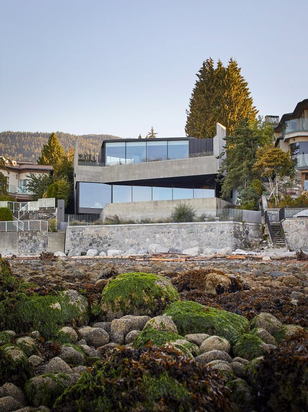 Exterior view of a modern multi-level waterfront residence set directly above a rocky shoreline. The house features broad floor-to-ceiling glazing, horizontal concrete bands, dark vertical screening, and landscaped terraces, with neighbouring homes and wooded hills in the background.